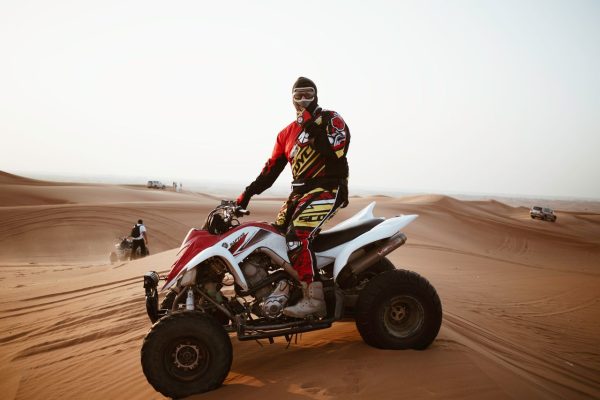 Full body of male biker in protective suit and helmet riding quad bike on sand in desert